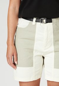 Helle Shorts mit grünen und cremefarbenen Panelen, ausgestattet mit einem schwarzen strukturierten Gürtel, seitlichen Taschen und einer lockeren Passform.