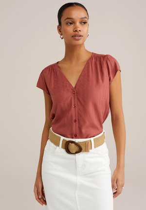 Blusa a maniche corte color ruggine con scollatura a V e motivo sottile, apertura con bottoni, abbinata a una gonna bianca e una cintura in tessuto intrecciato color beige.