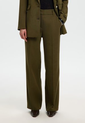Personne portant un pantalon large vert olive sur mesure et un blazer assorti avec des chaussures noires pointues.