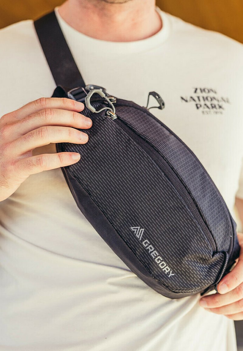 Black nylon sling bag with angled design, diagonal stripes, front zipper, and metal hardware. Logo branding visible on the side.