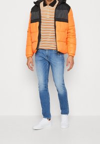 Man wearing orange and black puffer jacket, brown and white striped zip-up shirt, blue jeans, and white sneakers standing on white background.