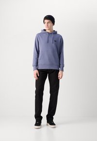Felpa in cotone viola con tasca a marsupio, cappuccio con cordino e un piccolo logo nero; abbinata a pantaloni neri e sneakers nere.