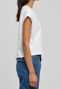 T-shirt bianco cropped con maniche arrotolate, realizzato in cotone. Abbinato a jeans di denim blu, in cui viene mostrata una vista laterale dell'outfit.
