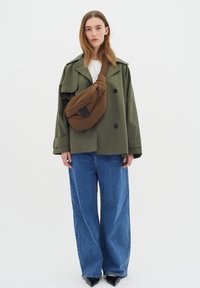 Trench coat verde sopra una camicia bianca, abbinato a jeans a gamba larga blu e una borsa a tracolla marrone. Scarpe nere a punta completano il look.