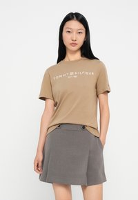 Καφέ κοντομάνικο μπλουζάκι με λευκό λογότυπο "TOMMY HILFIGER", συνδυασμένο με γκρι πλισέ φούστα που διαθέτει δύο μαύρα κουμπιά.