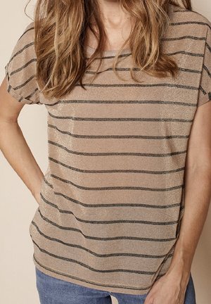 Mujer vistiendo una camisa beige de manga corta con delgadas rayas horizontales negras y jeans azules, de pie con una mano en la cadera.