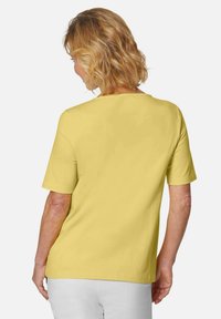 Geel T-shirt met korte mouwen en een eenvoudige ronde halslijn, gemaakt van een zachte stof, gedragen met witte broek, gezien van achteren.
