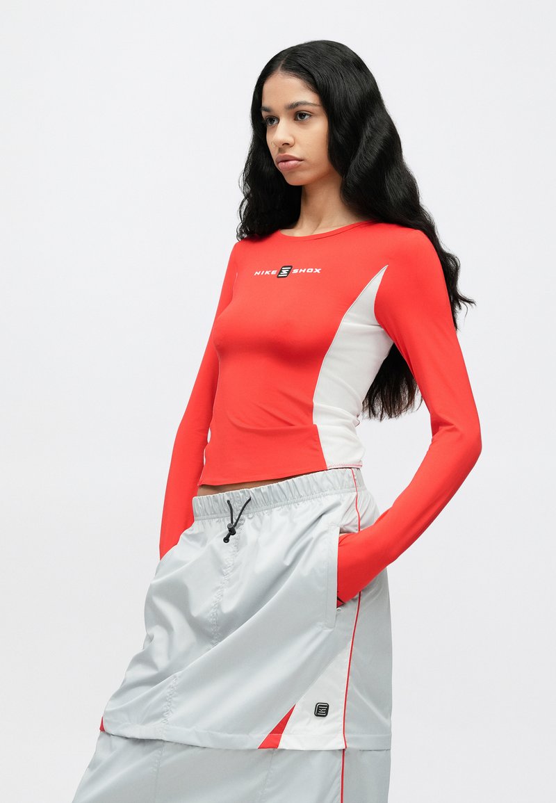 Rood top met lange mouwen met witte accenten en Nike Shox-logo; gecombineerd met losse grijze sportshorts met trekkoord en zijzakken.