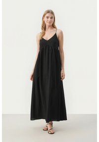 Juoda maxi suknelė su plonomis petnešėlėmis, V formos iškirpte, surinkta medžiaga krūtinėje ir lengva sijono dalimi. Modelis dėvi juodas šlepetes su dirželiu.