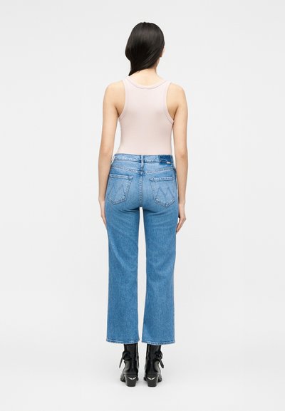 Γυναίκα που στέκεται με την πλάτη γυρισμένη, φορά ανοιχτόροζ αμάνικη μπλούζα, μπλε τζιν με ψηλή μέση και μαύρα μποτάκια σε λευκό φόντο.