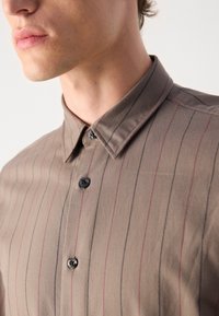 Camicia marrone con bottoni, caratterizzata da strisce verticali rosse e nere. Colletto classico, texture liscia e bottoni neri sul davanti.