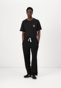 T-shirt noir avec un petit design graphique blanc sur la poitrine, assorti à un pantalon noir ample et des baskets noires avec des rayures blanches.