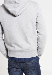 Felpa grigia in cotone con cappuccio regolabile, vista da dietro. Presenta un bordo e polsini a coste, abbinata a jeans blu scuro.