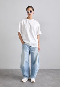 Tricou oversize din bumbac alb, blugi largi de culoare albastră deschisă, adidași albi și ochelari de soare cu nuanțe de violet, stând pe un fundal gri.