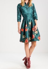 Teal button-up dress with floral pattern in reds and whites, 3/4 sleeves, cinched waist, and a flared skirt. Worn with pink ankle boots.