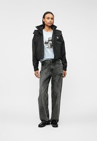 Μαύρο κομμένο μπουφάν με ψηλό γιακά, φορεμένο πάνω από ένα ανοιχτό μπλε μπλουζάκι με γραφικά. Συνδυάζεται με φαρδιά γκρι τζιν και μαύρα παπούτσια.