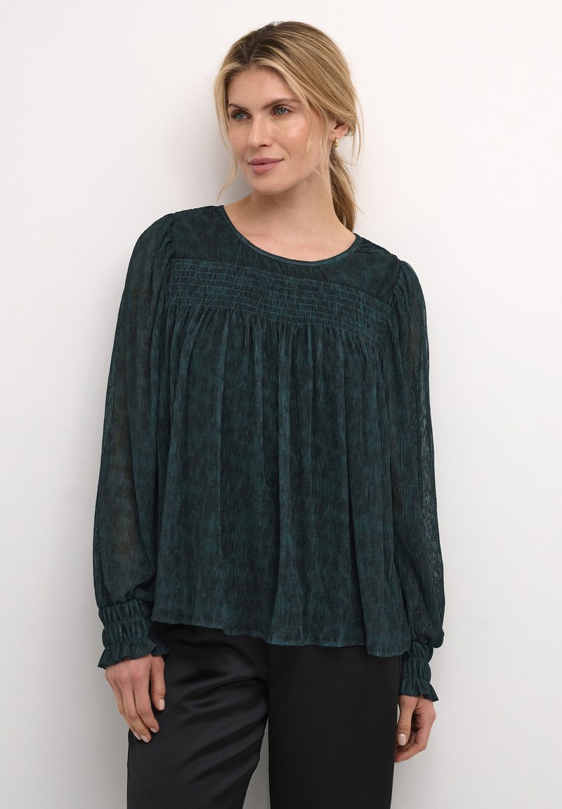 Blusa lunga teal a maniche lunghe con texture plissettata, caratterizzata da un corpino arricciato, scollatura a girocollo e polsini arricciati. Indossata con pantaloni neri.