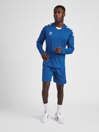 Blå atletisk trøje med lange ærmer, hvide detaljer og rund halsudskæring. Parret med matchende shorts. Båret med hvide atletiske sko.