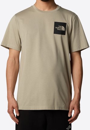 Camiseta beige de manga corta hecha de tela suave, con un parche de logo negro en el pecho izquierdo. Cuello redondo simple y ajuste holgado.