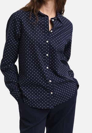 Kvinde iført navyblå skjorte med hvide polkaprikker og navyblå bukser, hænderne i lommerne, foran ensfarvet baggrund.