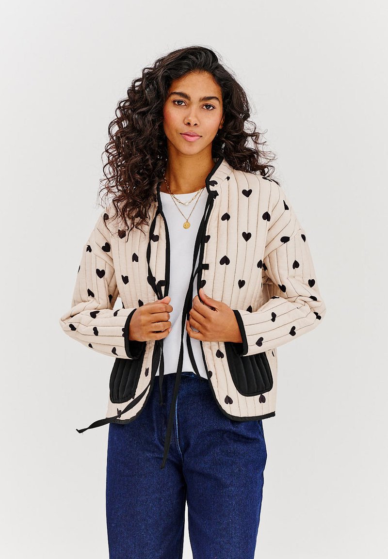 Prešívaná bomber bunda s krémovým základom, vertikálnymi pruhmi a čiernymi vzormi srdca. Obsahuje čierne panelové akcenty a predné šnurovanie.