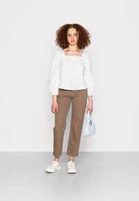 Blusa branca, à prova de ombros, com mangas bufantes, combinada com calças castanhas claras de perna reta e ténis brancos. Asegura uma bolsa azul clara.