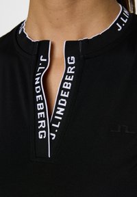 Svart pikétröja med halvdragkedja och vit märkeskrage. Tillverkad av ett mjukt, stretchigt material med subtil logotypdetalj.