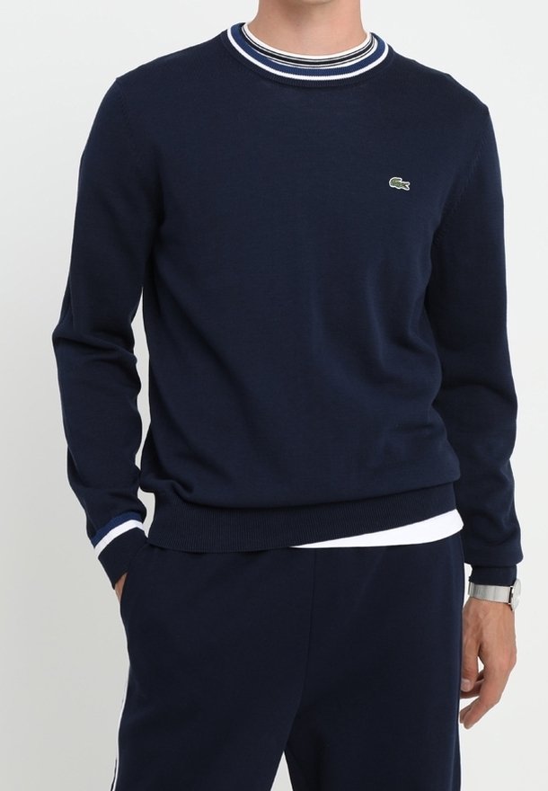 Homme portant un pull bleu marine à col rond avec un col et des poignets rayés blancs, assorti à un pantalon bleu marine et une montre-bracelet.
