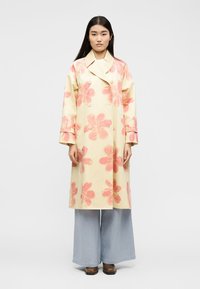 Γυναίκα που φοράει μακρύ παλτό σε κρεμ χρώμα με μεγάλα ροζ λουλουδάτα σχέδια, ανοιχτό μπλε παντελόνια με φαρδιά μπατζάκια και καφέ παπούτσια, στέκεται μπροστά σε λευκό φόντο.