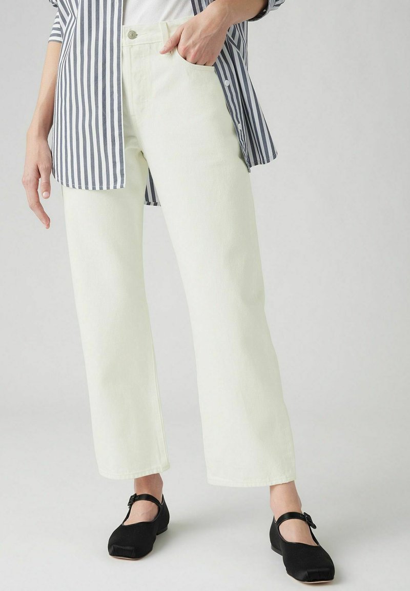 Lichtgele wide-leg jeans met een hoge taille, zijzakken en een klassieke knoopsluiting, gecombineerd met zwarte schoenen en een gestreept shirt.