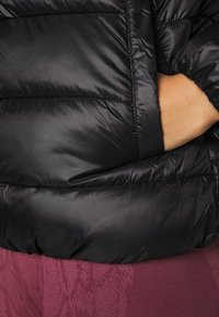 Svart puffjacka med ett kviltat mönster, med en glänsande och slät yta. Inkluderar en sidoficka och bärs över ett vinrött plagg.