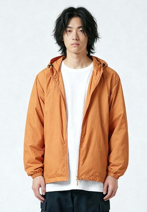 Giovane con capelli neri di media lunghezza che indossa una giacca con cappuccio arancione aperta sopra una maglietta bianca e pantaloni neri su uno sfondo semplice.