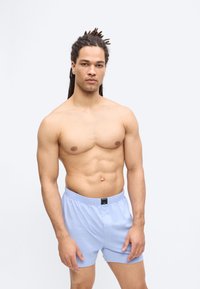 Homme musclé avec des dreadlocks portant un short de boxer bleu clair, debout et face à l'avant sur un fond blanc uni.