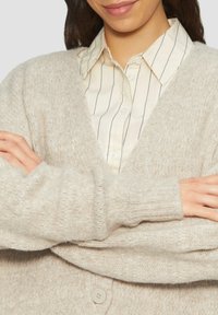 Beige gestrickte Strickjacke mit V-Ausschnitt, langen Ärmeln, gerippten Manschetten und einem einzigen Knopfverschluss, kombiniert mit einem cremefarbenen und marineblauen gestreiften Shirt.