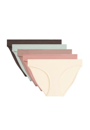 Femme portant un sous-vêtement style bikini en coton beige, montrée du milieu du torse aux cuisses supérieures sur un fond uni.
