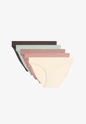 Femme portant un sous-vêtement style bikini en coton beige, montrée du milieu du torse aux cuisses supérieures sur un fond uni.