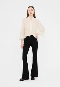 Bluză crem cu gât înalt și detaliu cu nasturi, asortată cu pantaloni evazați negri. Țesătură fină și croială ajustată. Pantofi maro.