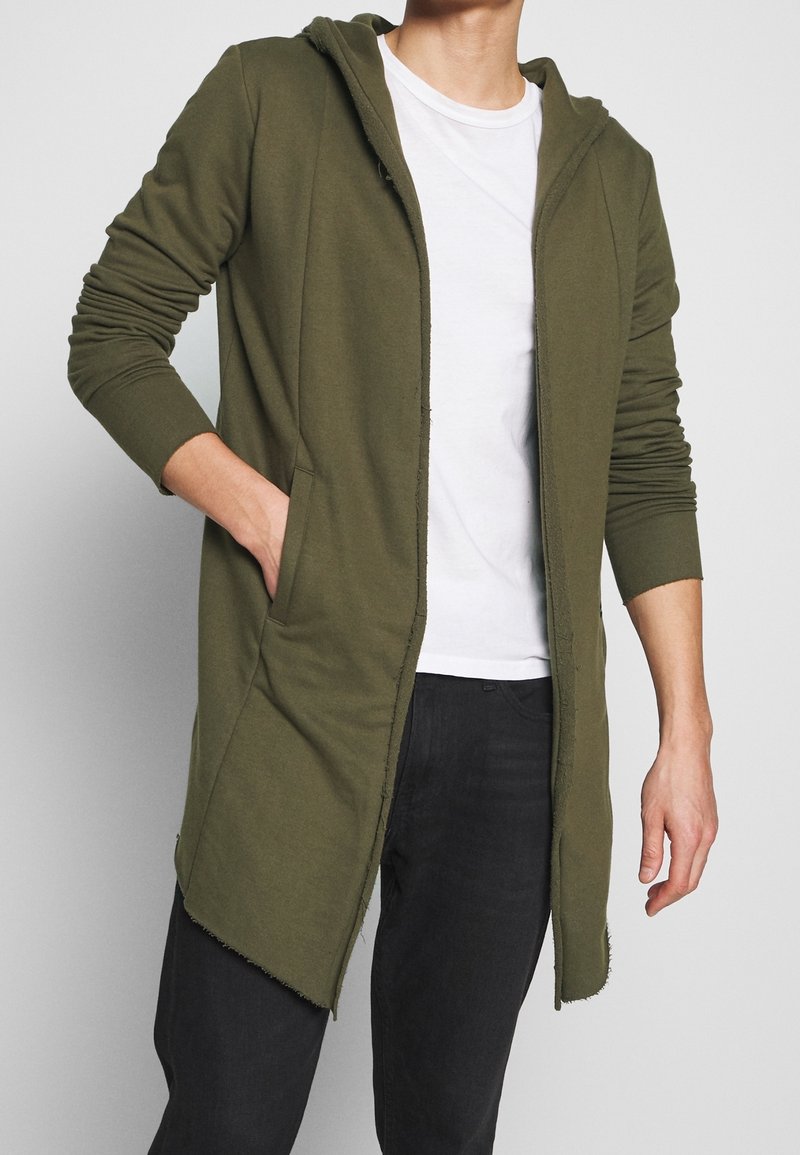Homme portant un sweat à capuche long ouvert de couleur vert olive avec poches, un t-shirt blanc et un pantalon noir, sur un fond clair uni.
