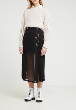 Femme portant un pull beige par-dessus une chemise blanche à col, une jupe midi noire avec des boutons dorés et des chaînes, ainsi que des bottines blanches à lacets.
