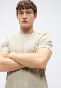 T-shirt desportiva bege com mangas curtas, tecido texturizado, gola redonda e pequeno logótipo no lado esquerdo. Ajuste elástico, design sem costuras.