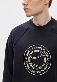 Tumši zils džemperis ar apaļu, bēša krāsas izšūtu logotipu "BOSS TENNIS CLUB" un tenisa grafiku, izgatavots no teksturētas auduma.