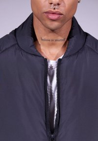 Casaco puffer preto com fecho de zíper e borda canelada, apresentando um colarinho alto. Camisa branca por baixo com um padrão subtil.
