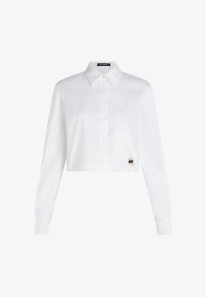 Cămașă albă scurtă cu mâneci lungi, cu butoniere în față, guler ascuțit și un buzunar pe partea stângă cu un mic design grafic alb-negru.