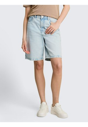 Osoba nosząca jasnoniebieskie dżinsowe spodenki do kolan, beżowy t-shirt i białe trampki, stojąca na jednolitym tle.