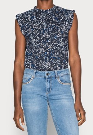 Woman wearing sleeveless navy blue floral top with ruffled shoulders tucked into light blue embellished jeans.