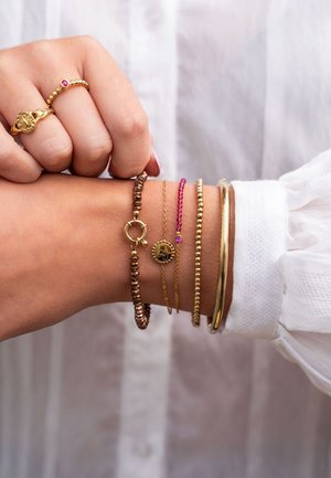Mano con cinco pulseras de oro y cuentas y dos anillos de oro, sobre un fondo de blusa blanca de manga larga.