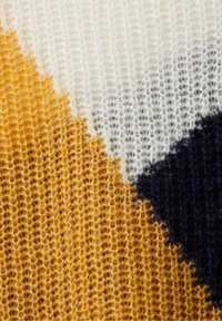 Detailní záběr na pleteninu ukazující texturovanou přízi v hořčicově žluté, bílé a tmavě námořnické modré barvě s geometrickým vzorem.