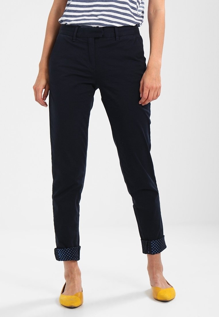 Pantalon bleu marine sur mesure en tissu de coton lisse, avec des revers roulés ornés d'un motif à pois. Porté avec un haut rayé et des chaussures jaunes.