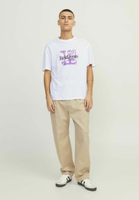 Hvid bomulds t-shirt med et lilla grafisk design og tekst, parret med beige bukser og hvide sneakers med sorte striber.