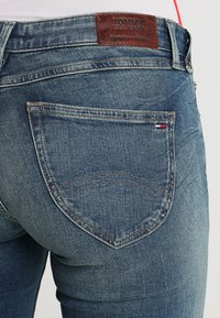 Jeans de mezclilla azul oscuro con una textura desgastada, que presentan un parche de cuero marrón con el logo de Tommy Jeans y bolsillos traseros con detalles de costura.